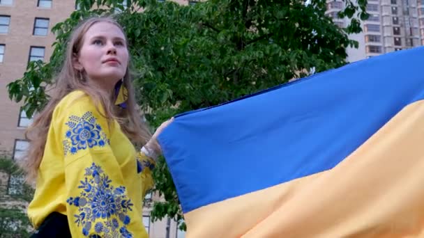 一个穿着绣花黄色衬衫 蓝色绣花的美丽的乌克兰女人举着乌克兰国旗 在房子的背景上 一棵树梦想着胜利的保护 请求帮助悲伤的眼睛 金黄的头发 — 图库视频影像