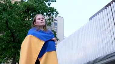 Genç kız, Ukrayna bayrağına sarılmış evlerin ve yeşil bir ağacın önünde. Zafer için bekliyor. Üzüntü, sarı saçlar. Geleceğe bak.