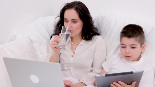 ガラスコップからきれいな水を飲む女性は 手でタブレットで4 5歳の息子の横にノートパソコンとベッドに座っているインターネット教育ゲームでオンライントレーニング息子と時間を過ごす — ストック動画