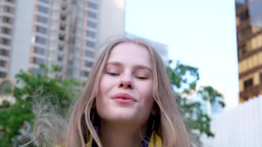 Sarı bluzlu genç kız dönüyor ve büyük bir şehirde yeşil bir ağacın arka planına hava öpücüğü yolluyor, sarı dalgalı saçlarıyla.