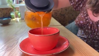 Çaydanlıktan alınan çay, açık pembe bir bardağa, tahta bir tabağın üzerine dökülüyor. Kahverengi elbiseli kız tanınmaz halde. Lezzetli içecekler vitaminlerden faydalanıyor.