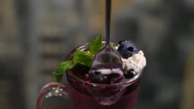 Cam bardaktan çay kaşığı naneli ve kremalı yaban mersinli tatlı almak. Karanlık bir arka planda insan eli var.