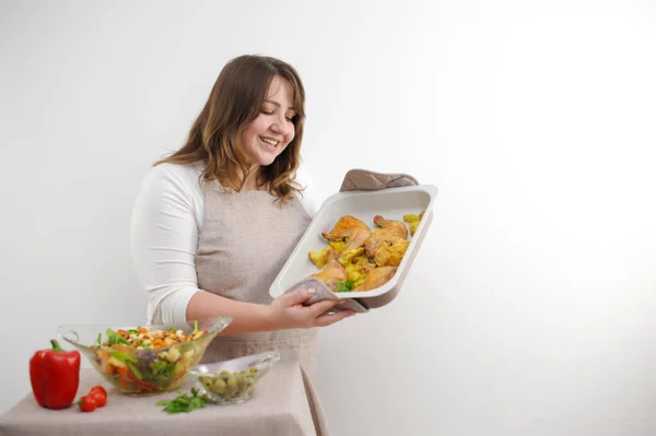 happy satisfied beautiful plump woman with a baking sheet in her hands delicious food chicken legs salad freshly prepared dinner for the family on a white background cook tasty laugh sincere facial