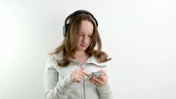 ヘッドフォンでプレイリスト10代の少女の適切な音楽のための電話で検索 白い背景で風スポーツウェアを聞くためのメロディを選択します — ストック動画