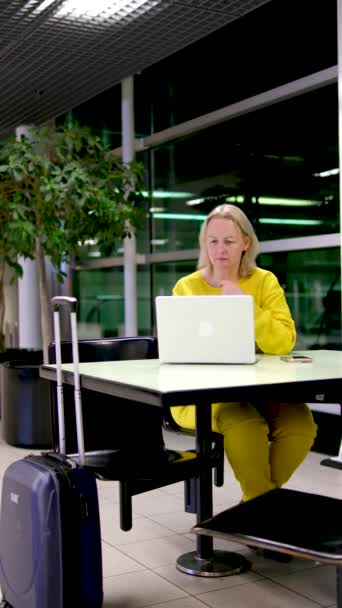 黄色スーツケースブルースーツケースで駅の女性作業ノートパソコンで待っています平面電車座っていますテーブルの上にもたれています彼女の腕の上に映画を見ます休憩オンラインインタビューの問題を解決する — ストック動画
