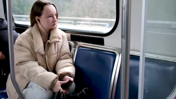 十代の女の子乗ります電車の中でバスヘッドフォンで耳の中の音楽に耳を傾ける秋の冬暖かいベージュのジャケット髪は下に縛られています本当の人座っている悲しい夢の下に隠された足待っています停止 — ストック動画