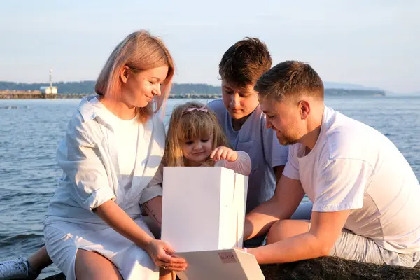 Happy family celebrating little girls birthday on the seashore mom brings a box with cake girl opens a gift dad mom brother and sister on the ocean shore. on a sunny day