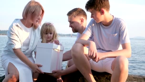 海岸で小さな女の子の誕生日を祝う幸せな家族は ケーキの女の子が海岸にギフトパパのお母さんと妹を開くボックスを持って来ます 晴れた日には — ストック動画