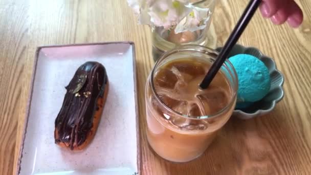 在杯子里搅拌海绵状的稻草 和奶油一起喝一杯美味的咖啡 在餐馆的木制桌子上放着蛋糕和蓝栗子 — 图库视频影像