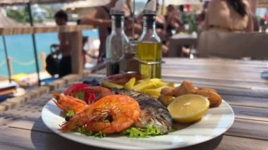 Arnavutluk 'un Ksemil tatil beldesindeki plaj şık restoranında bir kız bıçak ve çatalla balık yiyor ve genç bir kadın gölgede oturuyor büyük deniz mahsulleri ve Dorado balığı yiyor. Tatilde zaman geçirmek