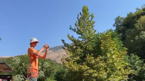 一位年轻的摄影师拍摄了一张阿尔巴尼亚标志性的蓝眼睛少年的照片 照片背景是身穿巴拿马帽和橙色夹克的天空和山脉阿尔巴尼亚09 2023 — 图库视频影像