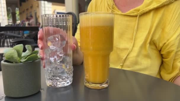 美味的橙汁 冰热的日子在灰色的桌子上女性的手把冰块倒入新鲜挤压的多果饮料慢动作视频小花盆 在科孚岛上服侍得真漂亮 — 图库视频影像