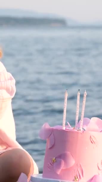 一个穿着粉红裙子的小女孩坐在那里 靠着美丽的大海 拿着三支蜡烛和一支钢笔 吃着一个粉红的蛋糕 可爱的小妖精斜视着 年幼的孩子们将来的生日 — 图库视频影像