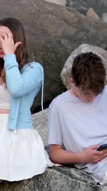 青少年们坐在石头上 男孩和女孩手里拿着手机 在初次约会时 他们使用的是依赖社交网络的小工具 青少年轻装 — 图库视频影像