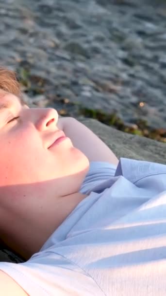 10代の少年は ビッグストーンショアのビーチに横たわっています 晴れた日 海の休息 頭の下に手を置くスニーカーのショートパンツ Tシャツの青年期の若者たちは — ストック動画