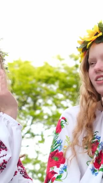 花輪を持つウクライナの女の子は 伝統的なウクライナの服に座っていると喜び 互いに耳の中に噂話をささやく興味深いことに聞く黒とブロンドの髪のガールフレンド民族衣装 — ストック動画