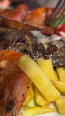 Arnavutluk 'un Ksemil tatil beldesindeki plaj şık restoranında bir kız bıçak ve çatalla balık yiyor ve genç bir kadın gölgede oturuyor büyük deniz mahsulleri ve Dorado balığı yiyor. Tatilde zaman geçirmek