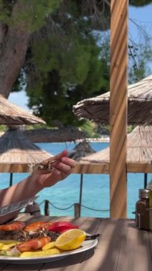 Arnavutluk 'un Ksemil tatil beldesindeki plaj şık restoranında bir kız bıçak ve çatalla balık yiyor ve genç bir kadın gölgede oturuyor büyük deniz mahsulleri ve Dorado balığı yiyor. Tatilde zaman geçirmek