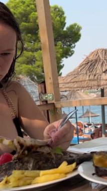 Plajdaki siyah mayo giymiş genç bir kız, Arnavutluk sahilinde Dorada balığı yiyor. Kızarmış sebzeleri beyaz tabakta kızartıyor. Öğle yemeğinde, güzel havalarda. seyahat