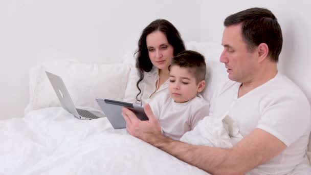 バナー 最新のテクノロジー家族の夫と子供は 一緒に白い服をリラックスしてベッド上のラップトップで映画を見ている オンライン親戚のお父さんと息子を笑って笑う笑顔 — ストック動画