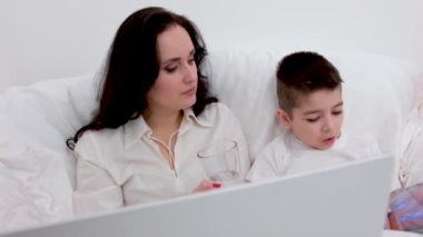 Anne ve oğlu battaniyenin altında 4-6 yaşlarındaki tabletçi çocukla oynuyorlar siyah saçlı güzel bir kadın beyaz çarşaf altında uzanmış gülücük oyunları internetine gülüyor. çevrimiçi öğrenim eğitim faaliyetleri