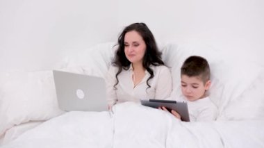 Anne ve oğlu battaniyenin altında 4-6 yaşlarındaki tabletçi çocukla oynuyorlar siyah saçlı güzel bir kadın beyaz çarşaf altında uzanmış gülücük oyunları internetine gülüyor. çevrimiçi öğrenim eğitim faaliyetleri