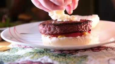 Kadın elleri ızgara hamburgerin üzerine rendelenmiş mozzarella peyniri serpiyor ketçaplı hardal tabağında ızgara hamburger pirzolası yiyor. gerçek zamanlı servis.