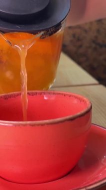 Çaydanlıktan alınan çay, açık pembe bir bardağa, tahta bir tabağın üzerine dökülüyor. Kahverengi elbiseli kız tanınmaz halde. Lezzetli içecekler vitaminlerden faydalanıyor.