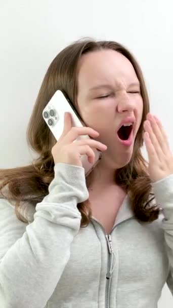 不愉快な退屈な会話を電話で話している間 若い女の子はスタジオで休むことに疲れている手のヨーンティーンエイジャーで口を覆います 白い携帯電話のコンテストは不満のマグに直面しています — ストック動画