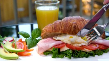 Kahvaltıda çatal ve bıçaklı yumurta ile kruvasan sandviçin üzerinde sarı sarısı akarsudaki akarsu tabaklarına akar. Avokado salatası kırmızı biberli turp kruvasanı bir sürü yeşillik doldurur. domates peyniri eti