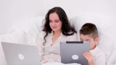 Anne ve oğlu beyaz battaniyenin altında yumuşak bir yatakta erkek tabletinin ellerinde sosyal ağlarla oynuyorlar. 4-6 yaşlarında kadın bilgisayarlı uzaktan kumanda konferansında serbest çalışıyor.