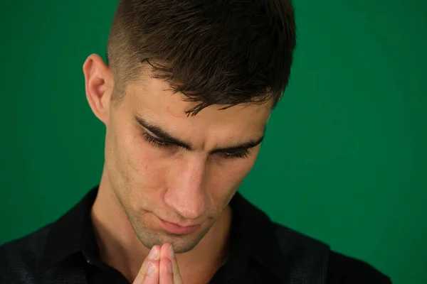 emotions of a handsome man guy on a green background chromakey close-up dark hair young man. man bowed his head praying to God on a black background at home. Holding hands in prayer, eyes closed.