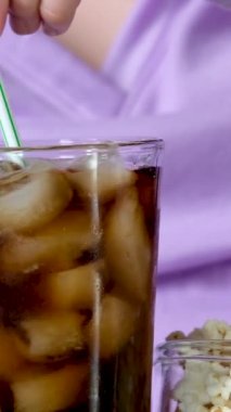 Yakın plan Coca-Cola bardağı ve cam tabağın yanında bol buzlu leylak rengi mor kazaklı patlamış mısırcı kız samanla içki karıştırıyor. 