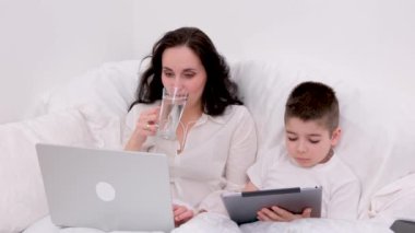 Cam bardaktan temiz su içen bir kadın, elinde tabletle dört-beş yaşındaki oğlunun yanında yatağında oturup oğluyla internet eğitimi görüyor.