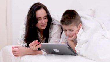 Anne ve oğlu battaniyenin altında 4-6 yaşlarındaki tablet çocuk üzerinde oynuyorlar siyah saçlı güzel bir kadın beyaz çarşaf altında yatıyor kahkaha atarak gülüyor internet üzerinden eğitim aktiviteleri öğreniyor