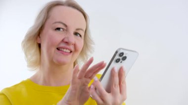 Telefondaki haberlere göz gezdirirken, beyaz arka plandaki parlak elbiseli yetişkin bir kadın, haberlere, sosyal ağlara bakıyor, reklam arıyor, satış, mesaj için yer arıyor, sarışın.