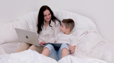 Çevrimiçi eğitim Apple iPad ve Macbook teknolojisini kullanarak yumuşak bir yatakta oturan bir kadın ve oğulun ellerinde rahat bir atmosferde oyun oynuyorlar. İnternetteki yaşam Ukrayna Vinnitsa 2023