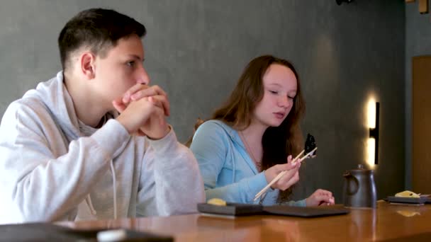 午餐或午餐在餐馆吃寿司的男孩十几岁的女孩在木桌边吃罗尔斯 旁边是灰色的墙 背景是筷子 是中国菜 是中国菜 青春期 — 图库视频影像