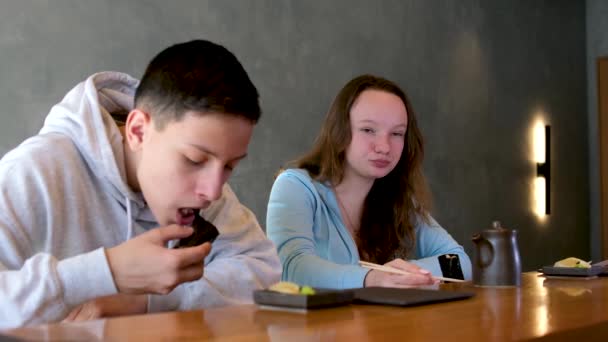 ランチやランチ レストランで寿司を食べる 10代の女の子は 箸中国料理の家族の兄弟姉妹との灰色の壁の背景に対してロールスロイスを食べます 青年期 — ストック動画