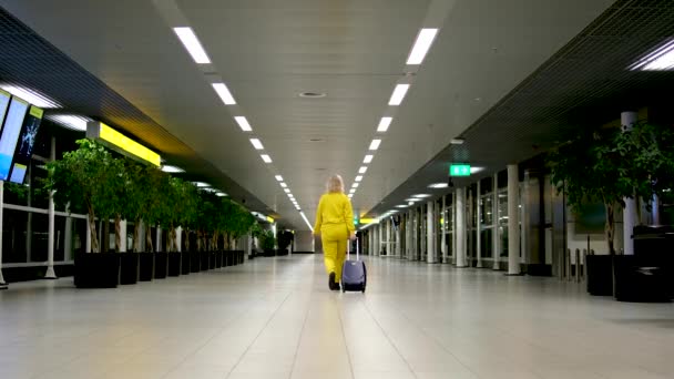 空港ターミナルでスーツケースを引っ張っている若い女性 窓の外で夜を待っているお別れのコミュニケーション挨拶を保持する黄色いスーツのスペースハンド荷物をコピーします — ストック動画