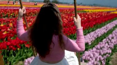 Bir salıncakta gezmek Avrupalı bir kızın pembe bluzunu geliştirir. Gökkuşağıyla bir çiçek bahçesinde oynayan beyaz etekli kız.
