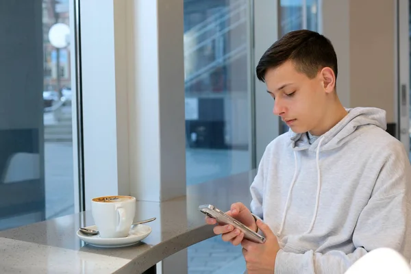 Sivilceli ergenlik çağında, telefonla kafede kahve içen, spor tişörtlü, camdan pencereli telefon internetindeki iyi wi-fi eğitimine bağlı beyaz koltuklu bir çocuk. Kişinin gerçek hayatı