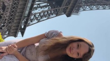 young beautiful girl in a blue dress sits near the Eiffel Tower near a green tree she straightens her braids looks around she is happy and very happy. High quality 4k footage