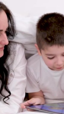 Anne ve oğlu battaniyenin altında 4-6 yaşlarındaki tabletçi çocukla oynuyorlar siyah saçlı güzel bir kadın beyaz çarşaf altında uzanmış gülücük oyunları internetine gülüyor. çevrimiçi öğrenim eğitim faaliyetleri