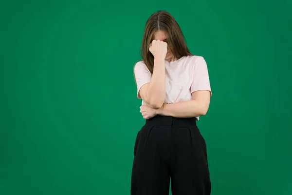 sad expression covering face with hands while crying. Depression concept. different emotions of a young girl on a green background chromakey beautiful facial features real people. pink T-shirt