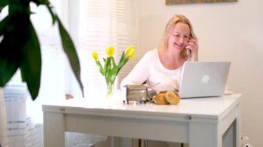 Orta yaşlı bir kadın akıllı telefon ve dizüstü bilgisayar kullanarak kahvaltı yapıyor, akıllı telefon uygulamalarını okuyup video izlemek için kullanıyor, kapatıyor. Yüksek kalite 4k görüntü