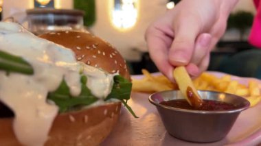 Patates kızartmasını arka planda hamburgerle ketçapın içine daldırırken yakın çekim. Kırmızı kamerayla çekilmiş. Yüksek kalite 4k görüntü