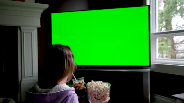 静态三脚架拍摄的年轻女子在电视绿色屏幕上看电影和吃爆米花而坐在沙发上 女孩们在家里客厅里观看彩色电视上她最喜欢的节目 — 图库视频影像