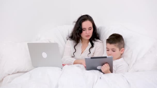 ホワイトブランケットの下のお母さんと息子は オンラインでソーシャルネットワークをプレイ少年タブレットの手の中に柔らかいベッドの上に座っている彼は4 6歳の女性はノートパソコンのリモートワークオンライン会議でフリーランスとして働いています — ストック動画