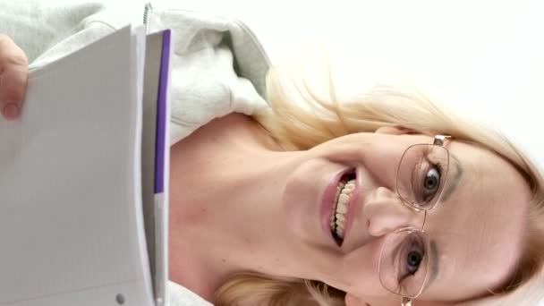 明るい背景に白いノートパッドを読んでブロンド笑うメガネを持つ親指の女性 誠実な笑顔は アイデアを書く 興味のある広告の提案を思いつきます 本を書く — ストック動画